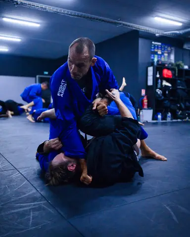 Un coach en kimono bleu Fight Arena contrôle son partenaire au sol pendant un échange de JJB